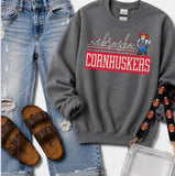 A gray crewneck sweatshirt featuring a 'nebraska CORNHUSKERS' graphic with the Cornhuskers mascot, styled in a flat lay with distressed blue jeans, brown slide sandals, and a clear handbag.